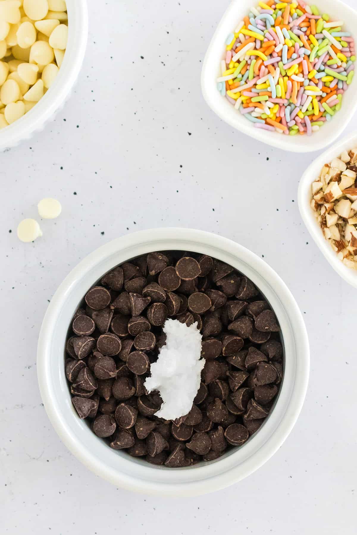 A bowl of chocolate chips with a dollop of coconut oil, surrounded by bowls of white chocolate chips, chopped nuts, and colorful sprinkles—perfect toppings for making delicious frozen chocolate bananas on a white surface.