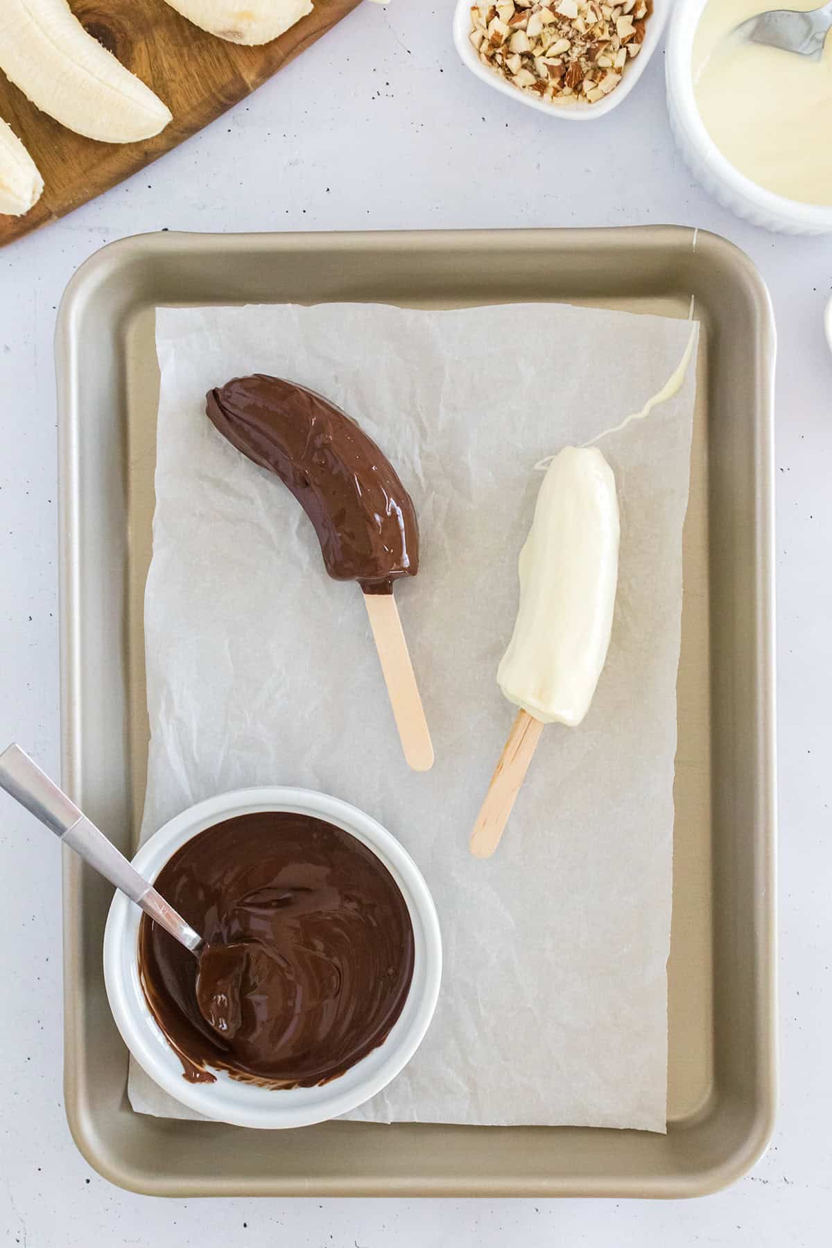 Two frozen chocolate covered bananas on sticks, one dipped in chocolate and the other in white chocolate, rest on parchment paper. Nearby, a bowl of melted chocolate with a spoon waits for more frozen bananas to be coated.
