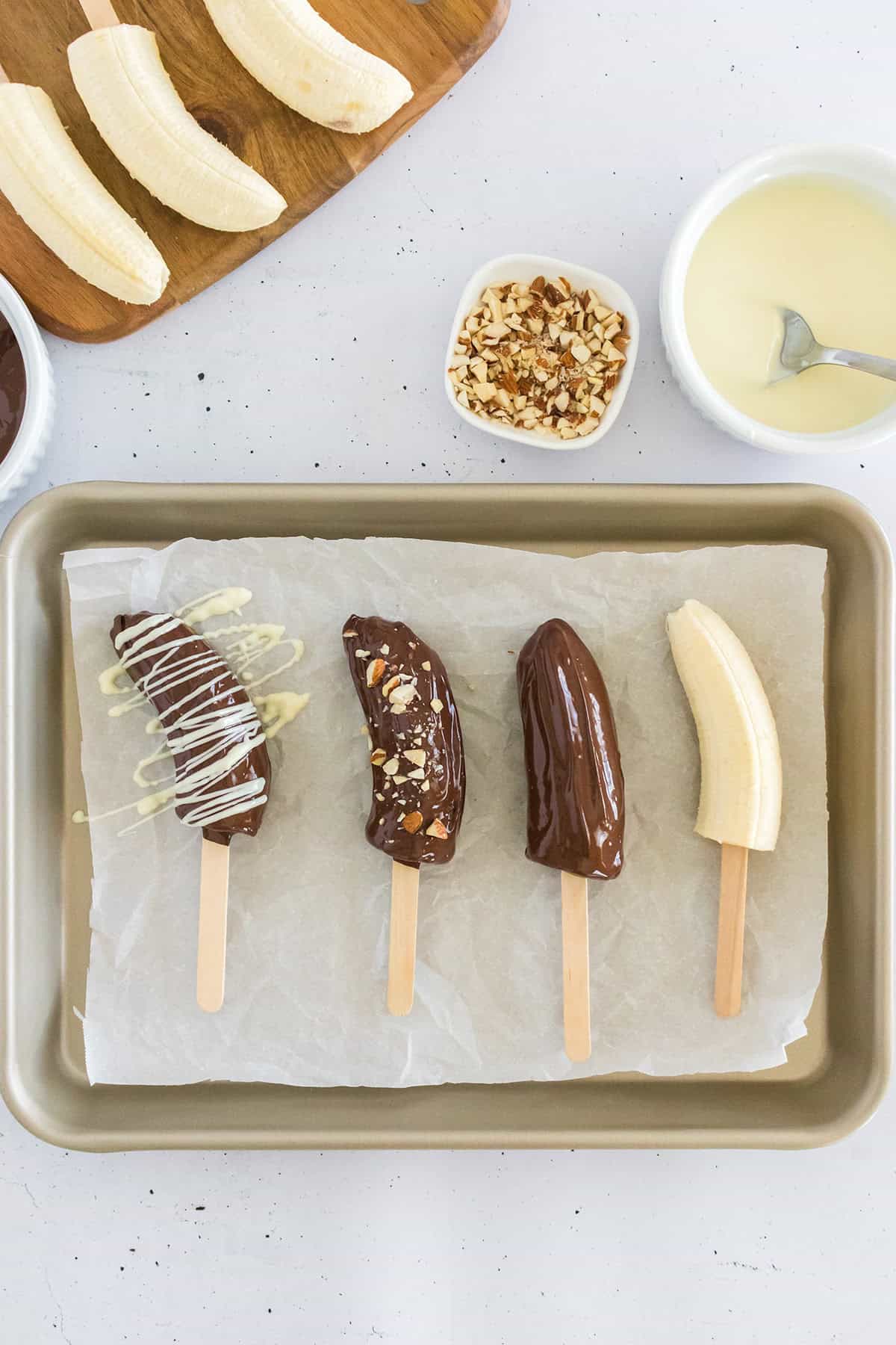 A baking tray with four frozen bananas on sticks: one plain, one chocolate covered, one with chocolate and nuts, and one drizzled with white chocolate. Ingredients for these frozen chocolate covered bananas are arranged nearby.