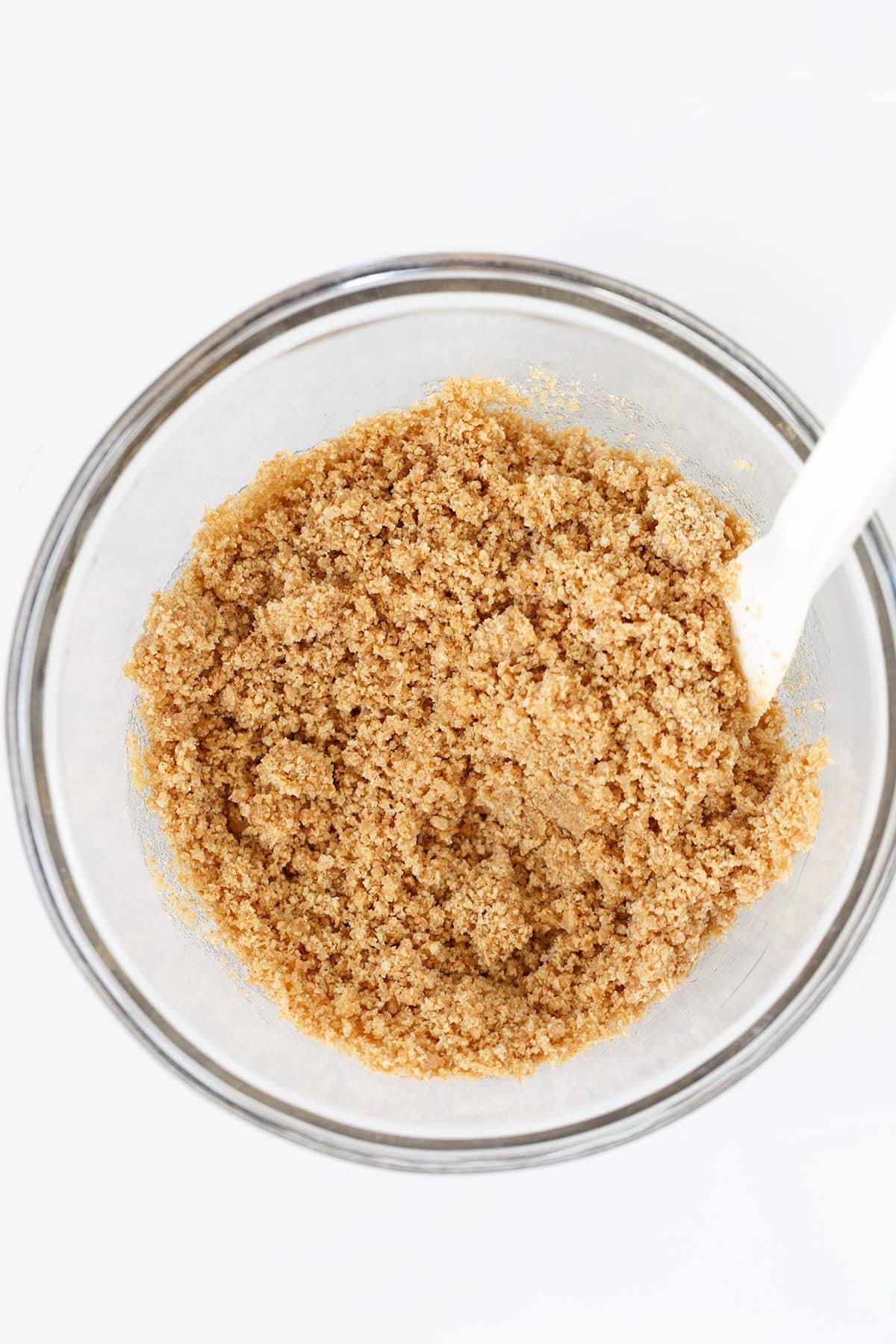 A glass bowl filled with a crumbly, light brown Graham Cracker Crust mixture and a white spatula on a white background.