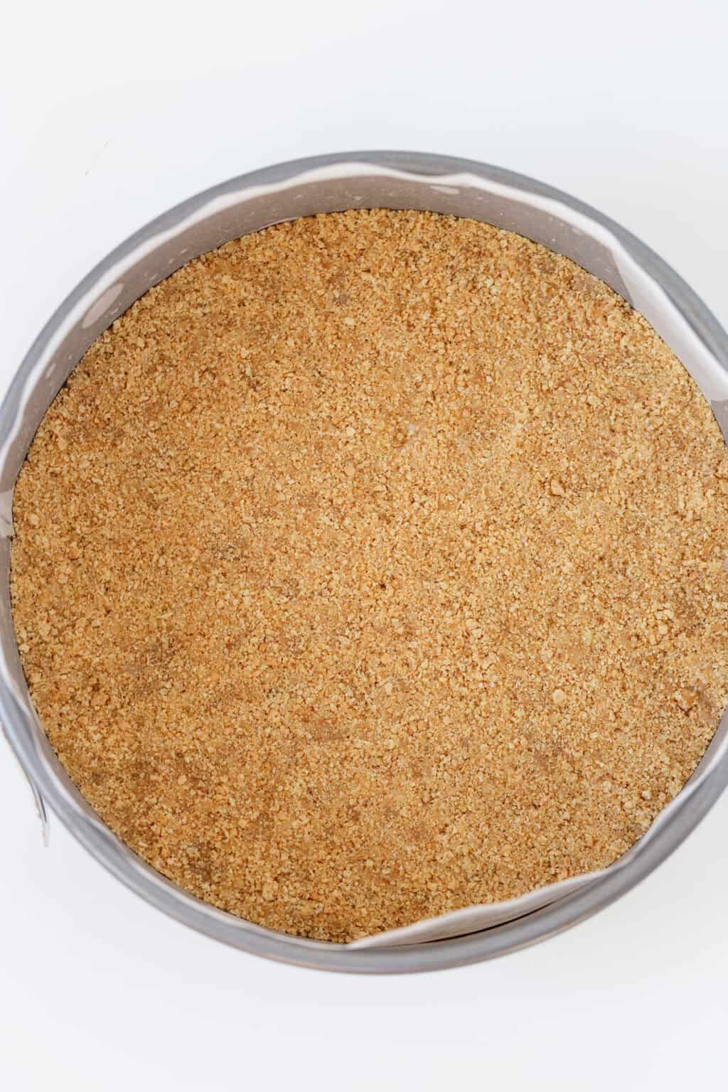 A round cake pan lined with parchment paper is filled with an even layer of homemade graham cracker crust, perfect as a base for your favorite pie crust or graham cracker crust recipe.