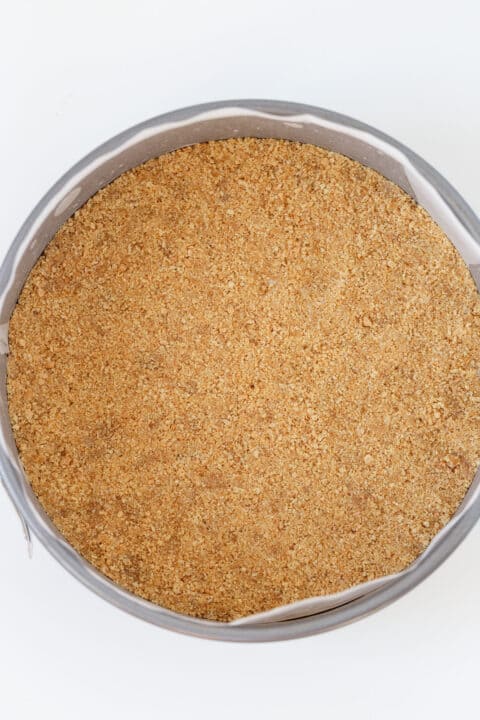 A round cake pan lined with parchment paper is filled with an even layer of homemade graham cracker crust, perfect as a base for your favorite pie crust or graham cracker crust recipe.