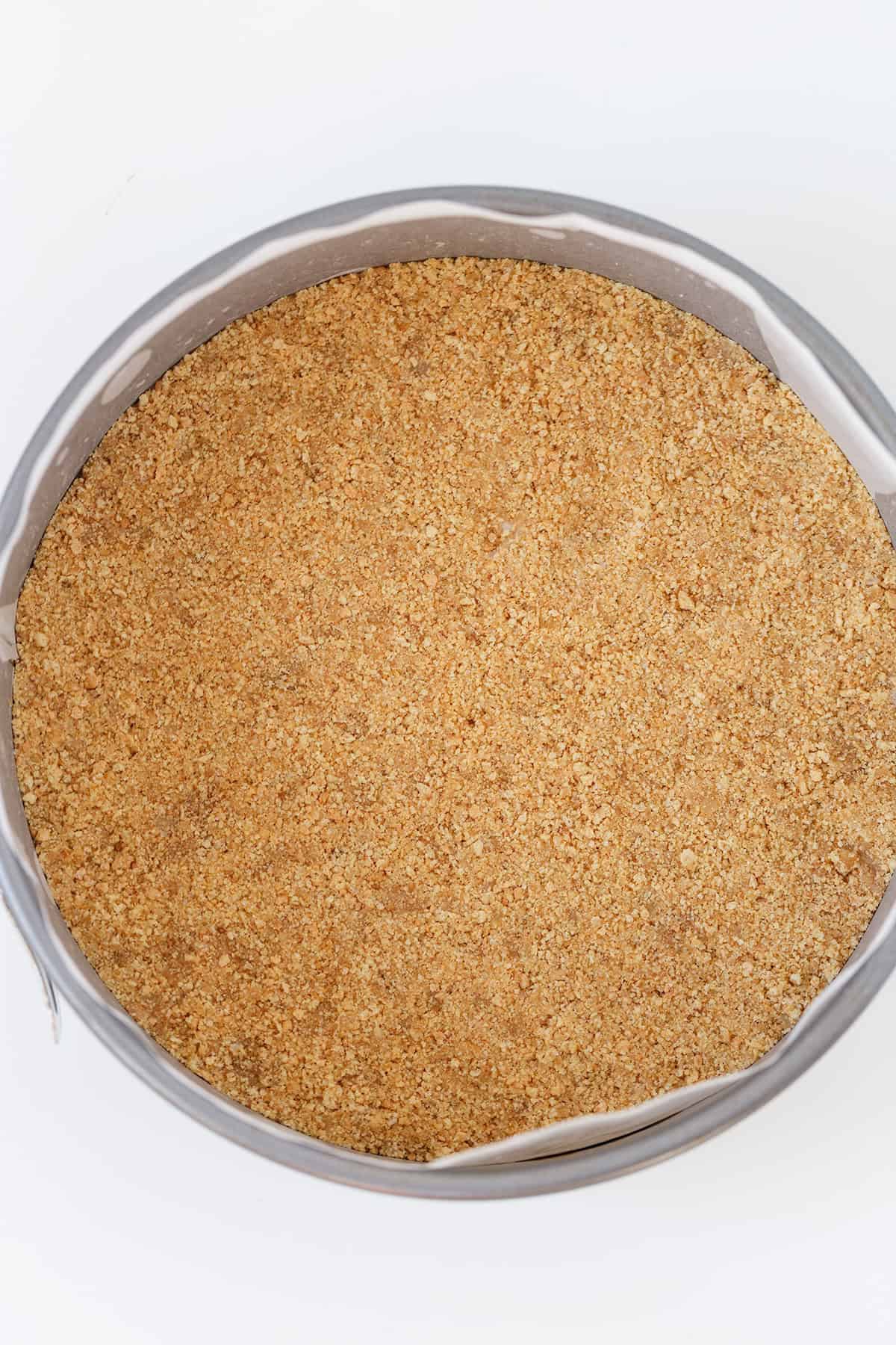A round baking pan filled with an even layer of crushed graham cracker crust, perfect as the base for a delicious Honey Bun Cheesecake, set against a white background.