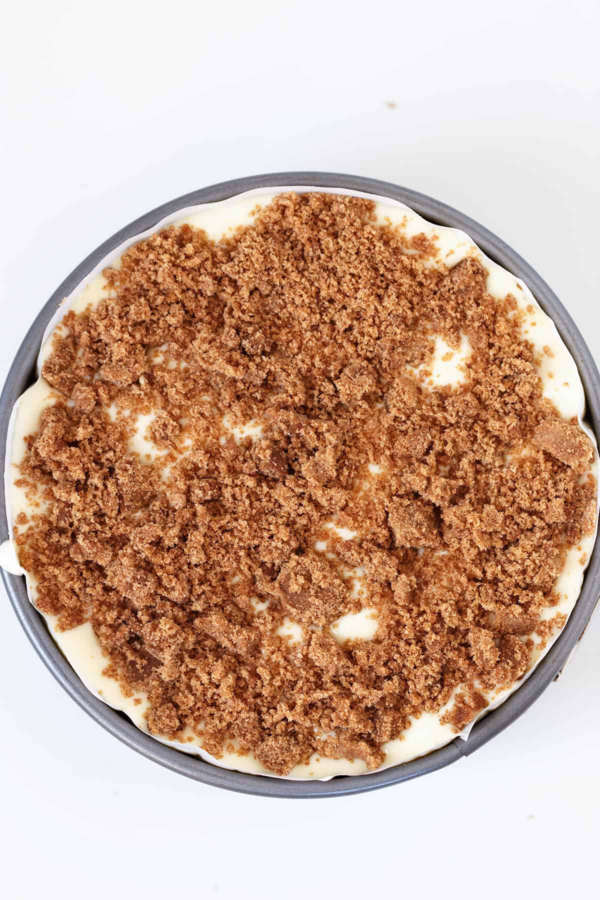 A round Honey Bun Cheesecake topped with a thick layer of crumbled brown sugar mixture sits on a white surface.