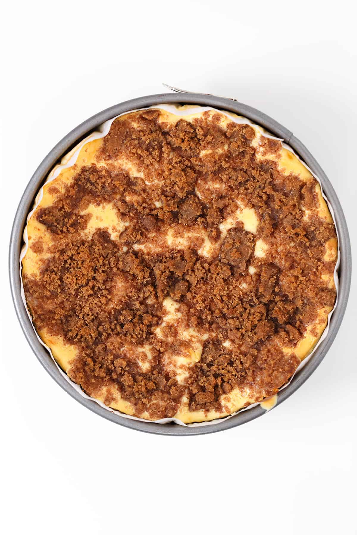 A round Honey Bun Cheesecake in a metal pan topped with a thick layer of brown crumb topping, photographed from above on a white background.