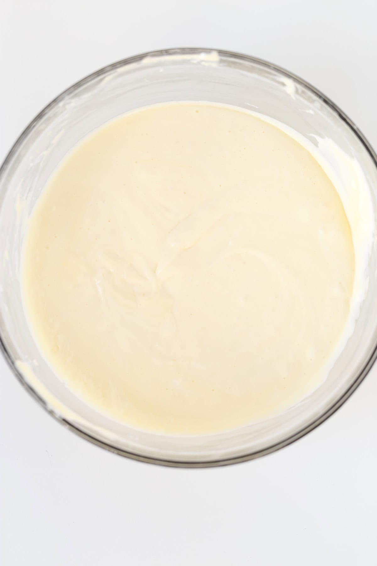 A clear glass bowl filled with smooth, pale yellow Honey Bun Cheesecake batter rests on a white surface.