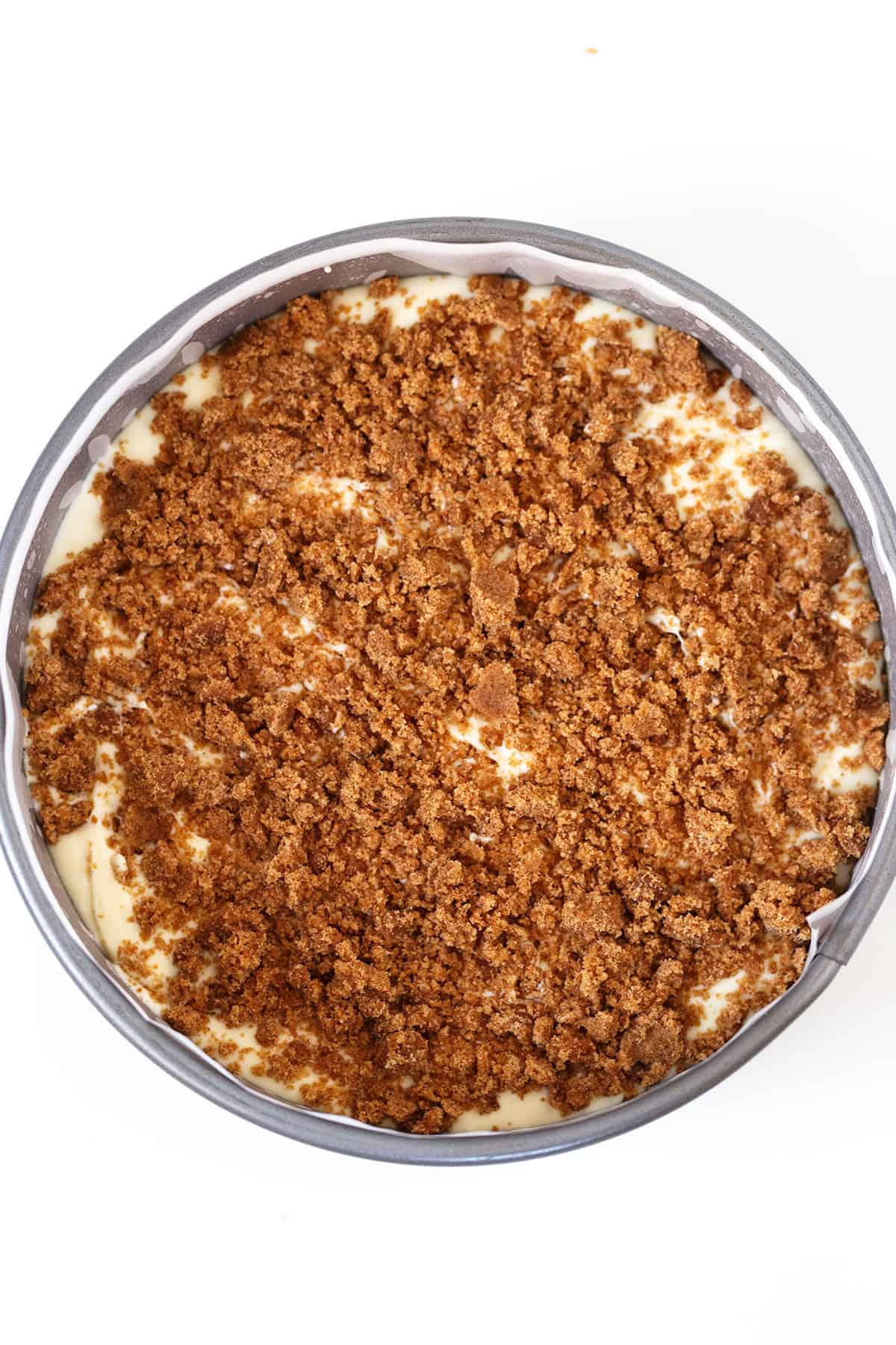 A round baking pan filled with a dessert topped with an even layer of brown crumbly streusel over a creamy Honey Bun Cheesecake base.