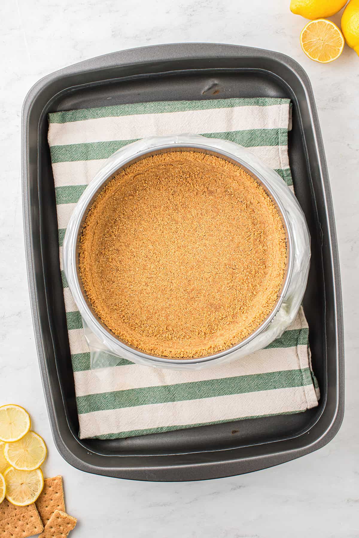 A round Graham Cracker Crust in a springform pan rests on a striped towel inside a baking tray, with graham crackers and lemon slices nearby, showcasing an inviting start to any pie crust recipe.