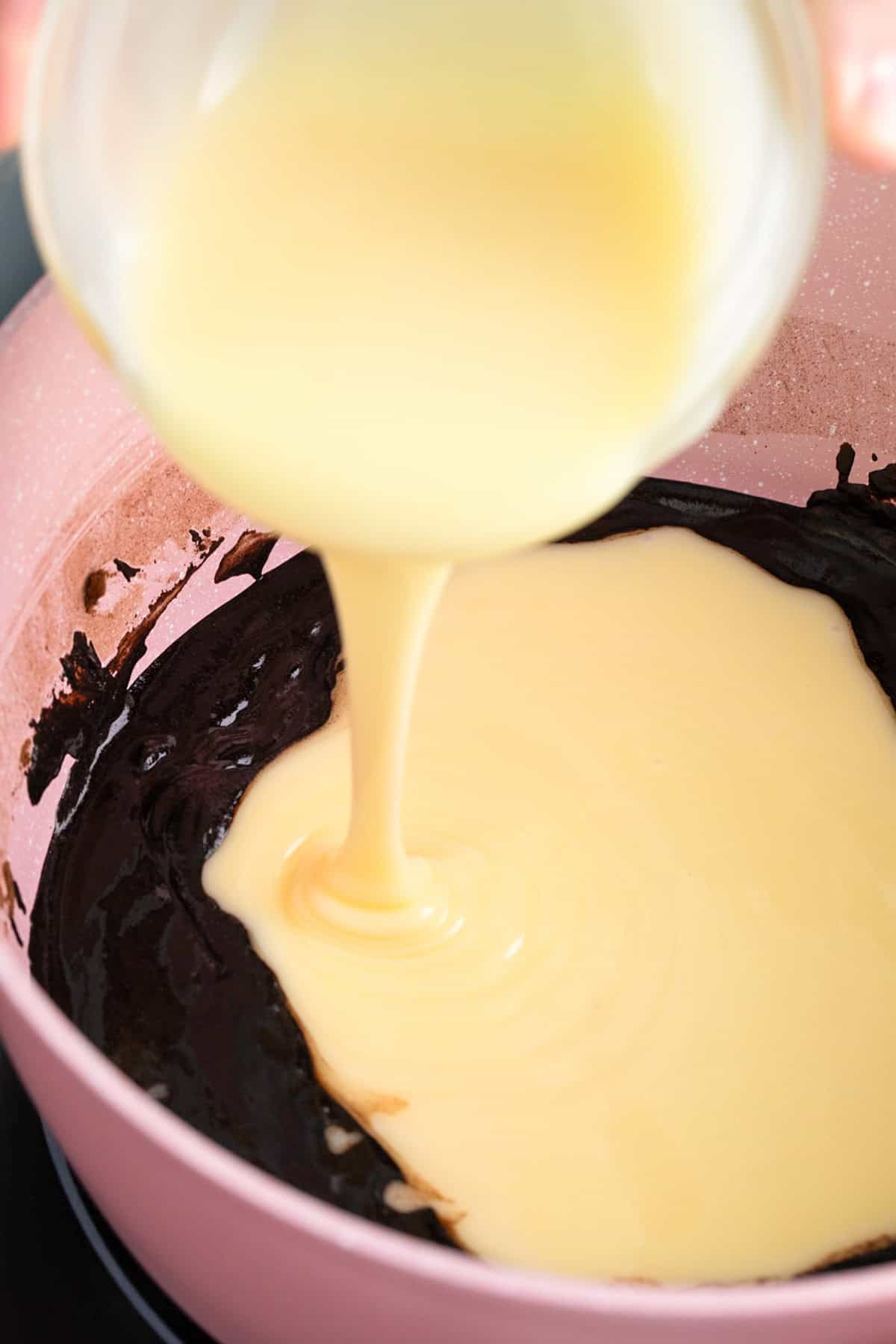 Sweetened condensed milk is being poured from a glass bowl into a saucepan filled with melted chocolate mixture, creating the gooey base for homemade Star Crunch Cookies.