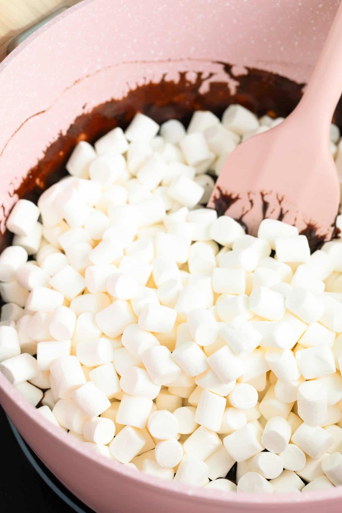 Mini marshmallows in a pink pot being stirred with a pink spatula, partially mixed with melted chocolate, perfect for making delicious Star Crunch Cookies.