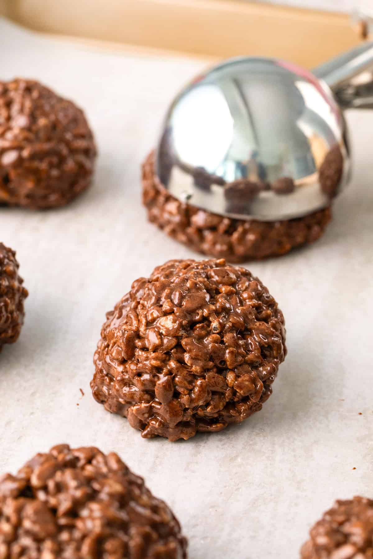 A metal scoop places a mound of chocolate rice cereal mixture onto parchment paper, forming Star Crunch Cookies, with several similar treats in the background.