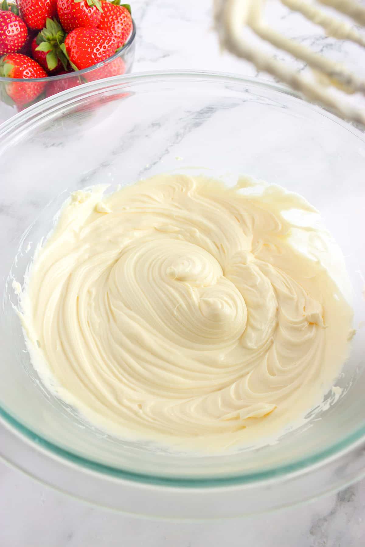 A glass bowl filled with smooth, creamy mixture sits on a marble surface, reminiscent of the perfect base for Strawberry Pizza, with a bowl of strawberries and a hand mixer in the background.
