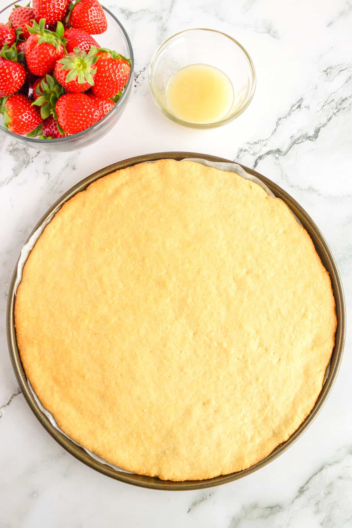 A round baked crust on a pizza pan, ready to become a delicious Strawberry Pizza, sits beside a bowl of strawberries and a small bowl of light-colored liquid on a marble surface.