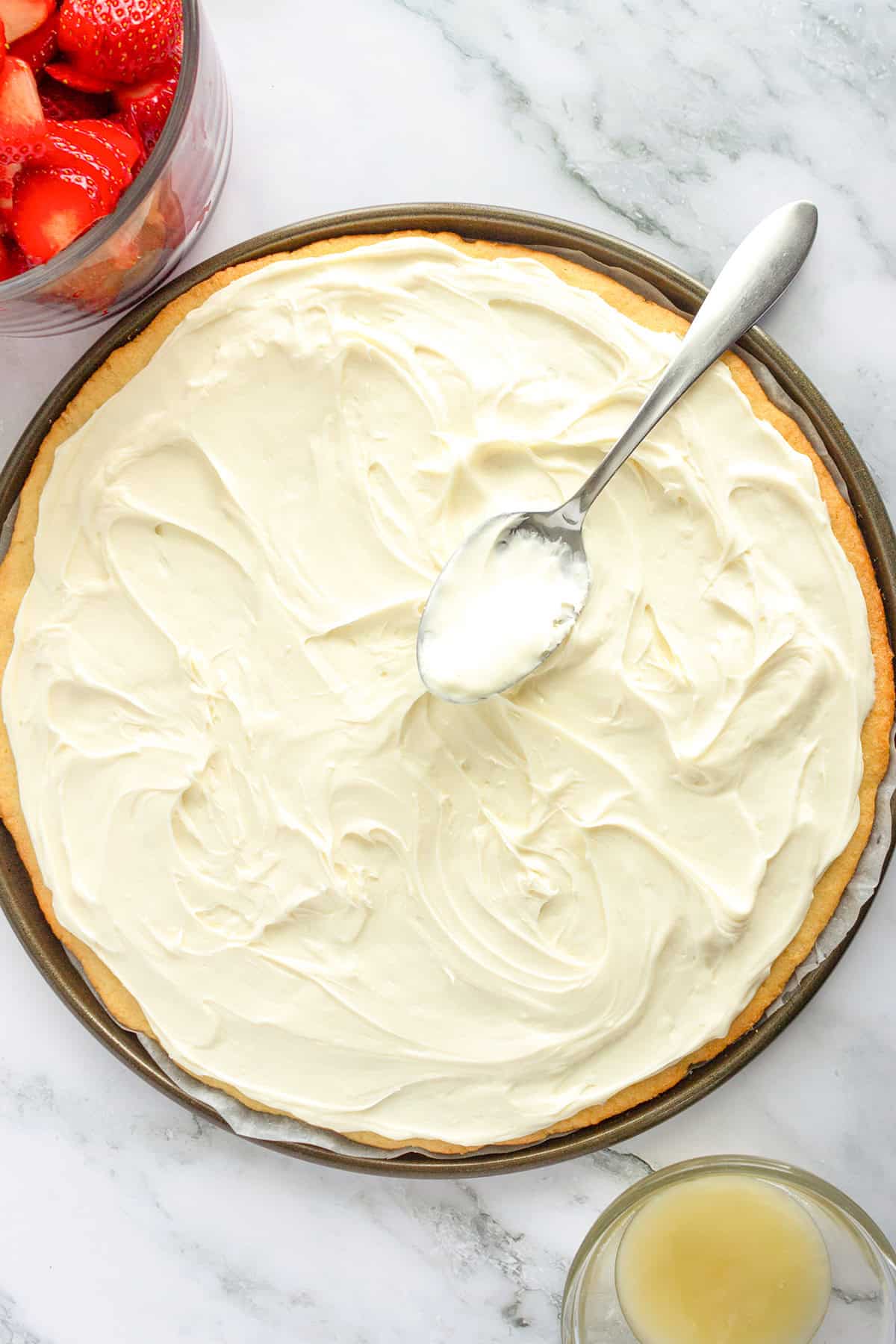 A round crust is topped with creamy white frosting, creating a tempting Strawberry Pizza. A spoon rests on the frosting, while sliced strawberries and a bowl of pale liquid sit nearby.