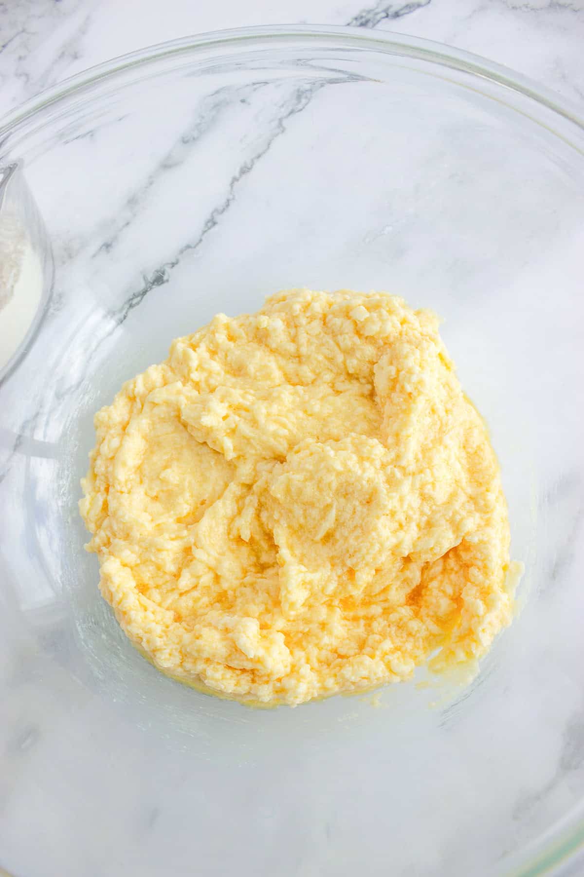 A glass bowl containing a mixture of creamed butter and sugar on a white marble surface, ready as the base for a delicious Strawberry Pizza.