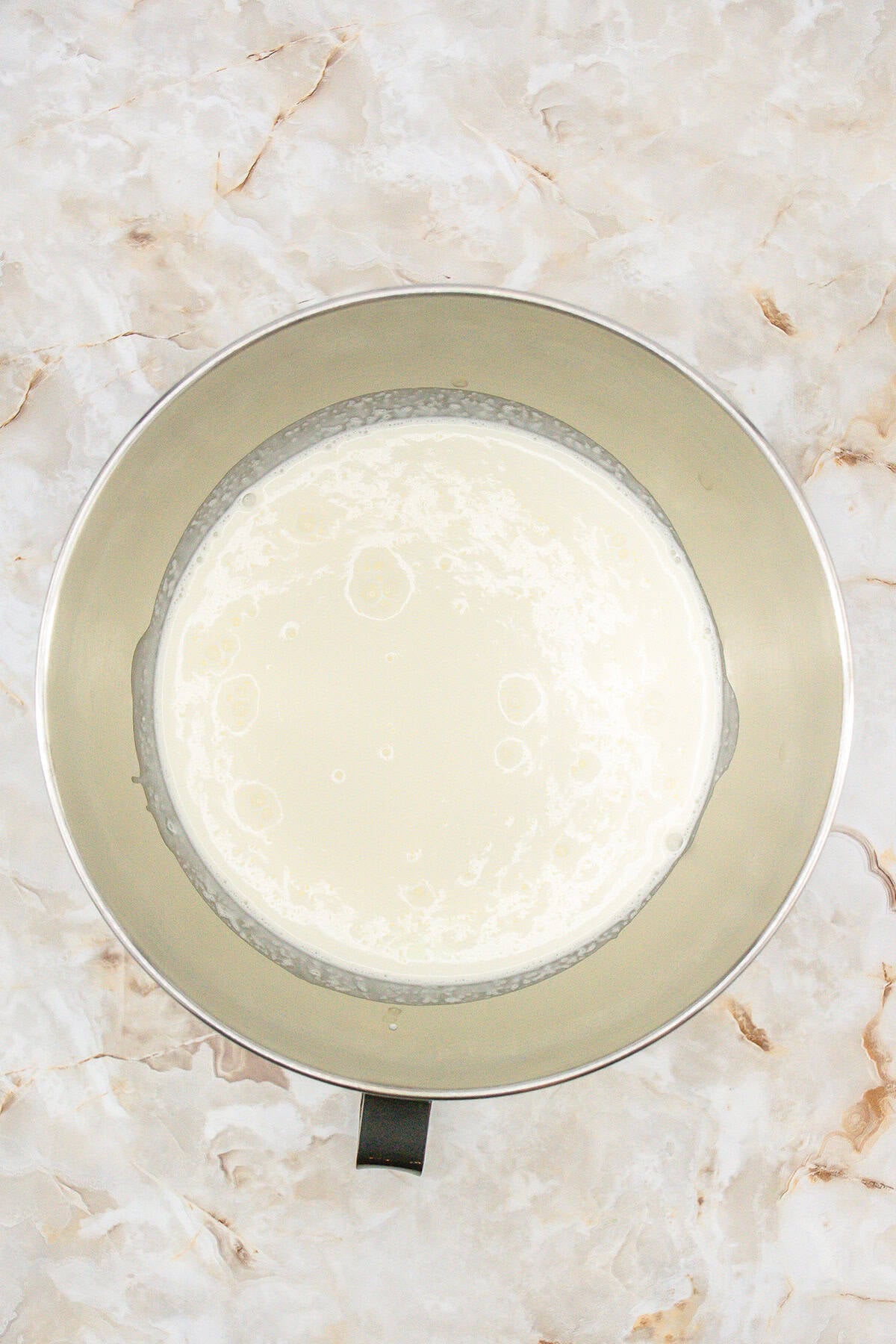 A metal mixing bowl containing milk for no churn vanilla ice cream is placed on a marbled countertop.