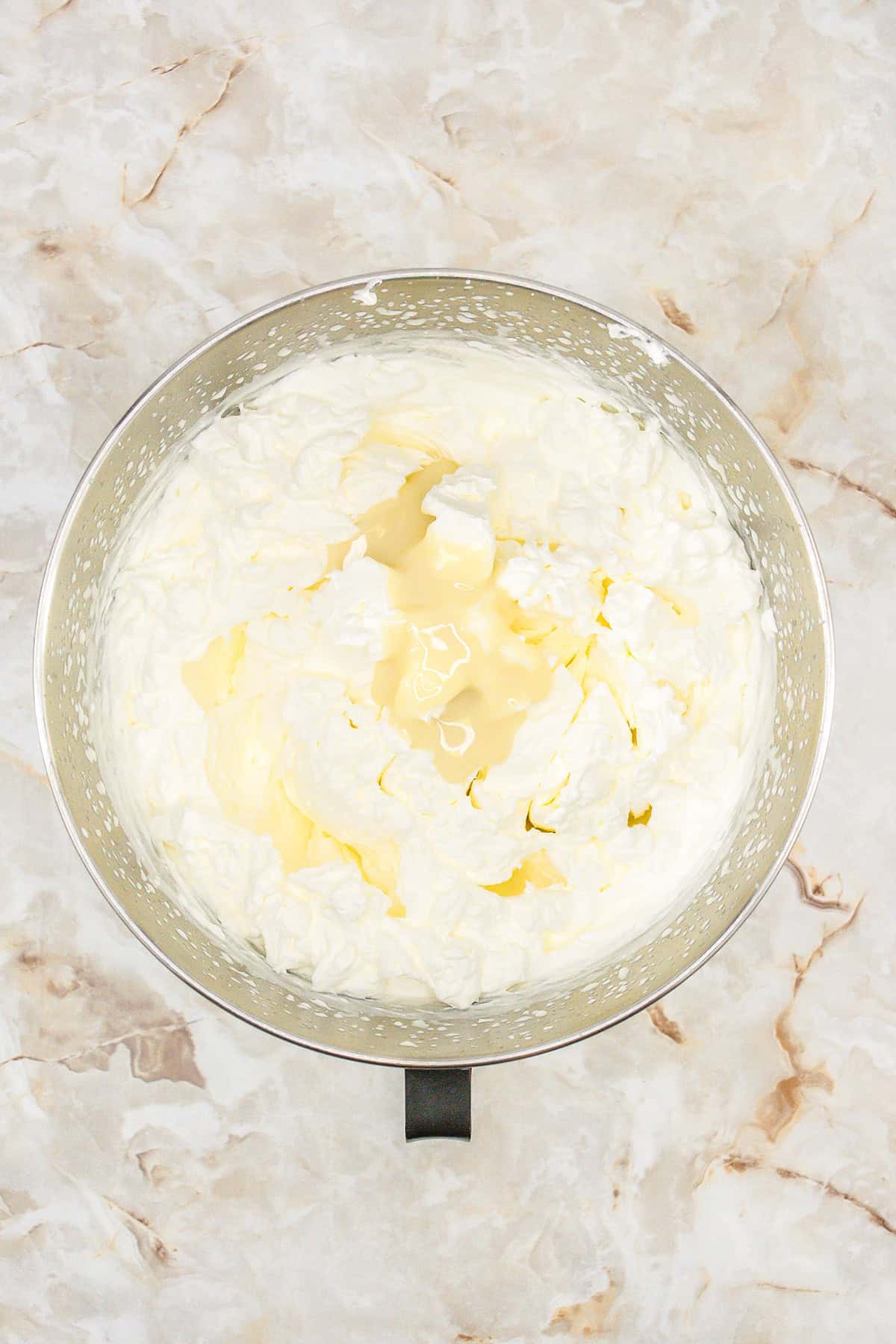 Metal mixing bowl filled with whipped cream on a light marble countertop, perfect for making no churn vanilla ice cream, viewed from above.