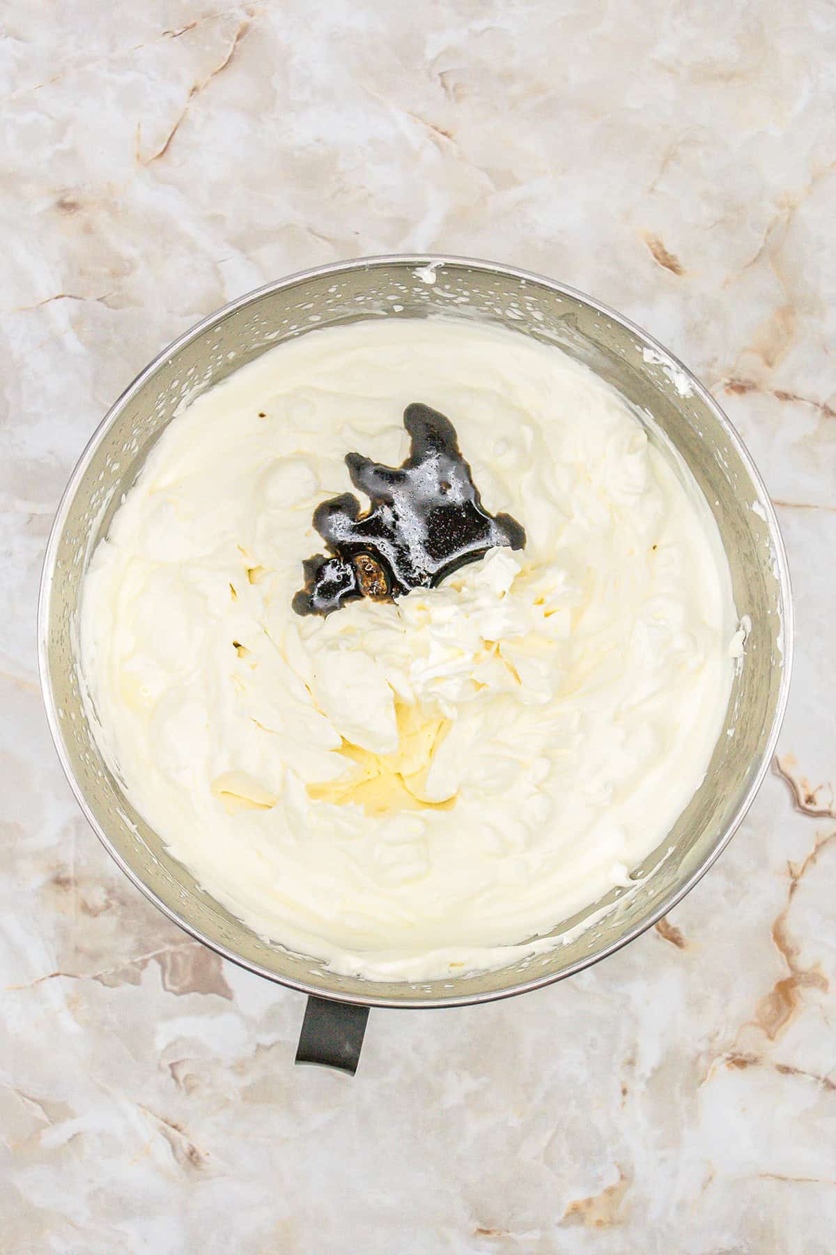 A metal mixing bowl filled with whipped cream and a dark liquid ingredient, possibly vanilla extract, on a marble surface—perfect for making easy No Churn Vanilla Ice Cream.