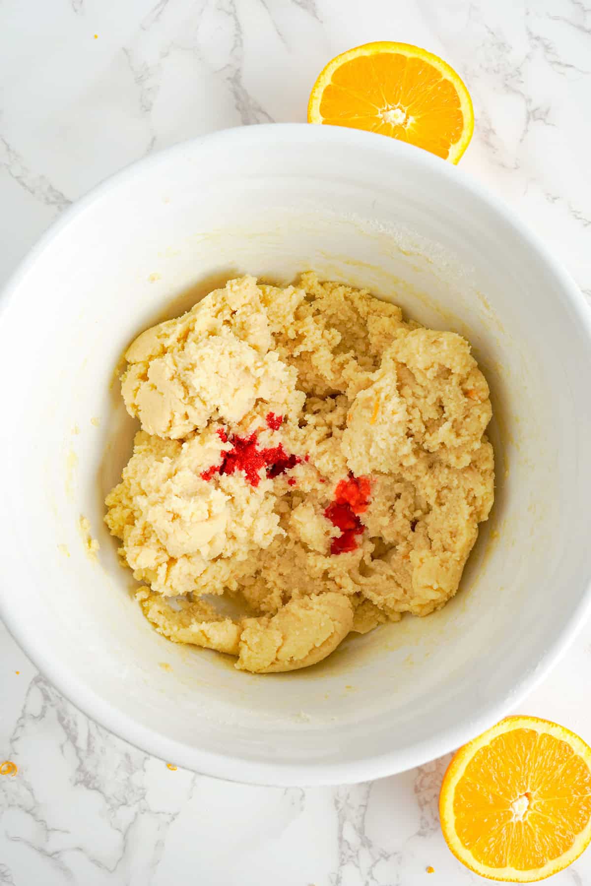 A white bowl filled with cookie dough and a few drops of red food coloring, perfect for making creamy cookies, with two orange halves placed beside the bowl on a marble surface.