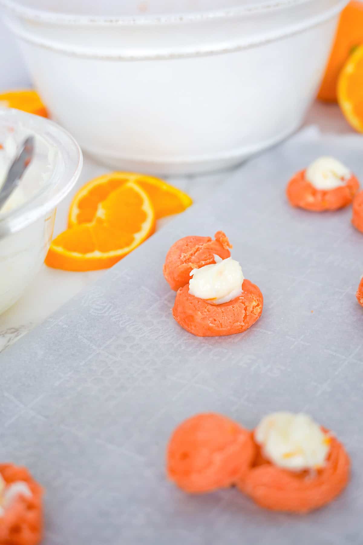 Orange creamsicle dough with white filling are arranged on parchment paper, resembling Creamsicle Cookies, with orange slices and mixing bowls in the background.