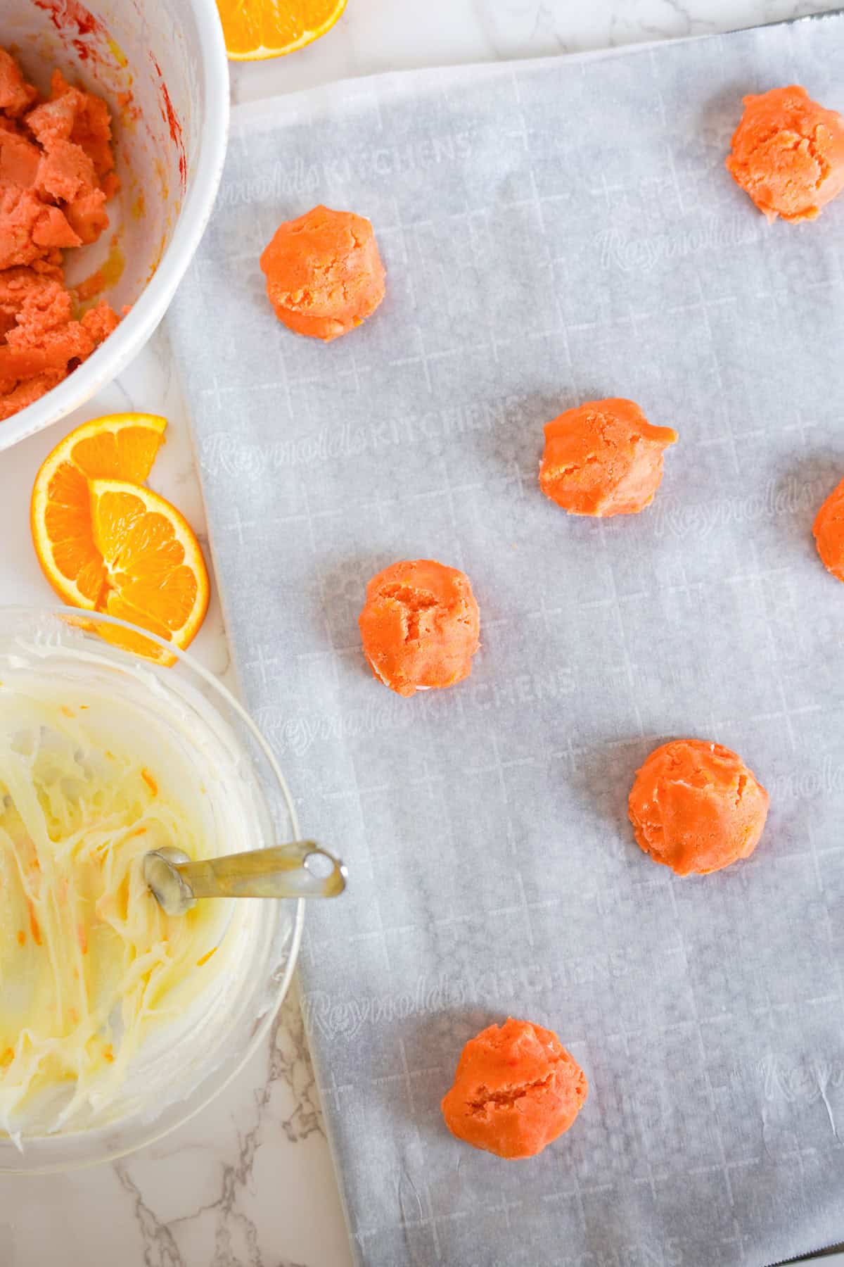 Round scoops of orange creamsicle cookie dough are arranged on a parchment-lined baking sheet, with orange slices and mixing bowls nearby on a marble counter.