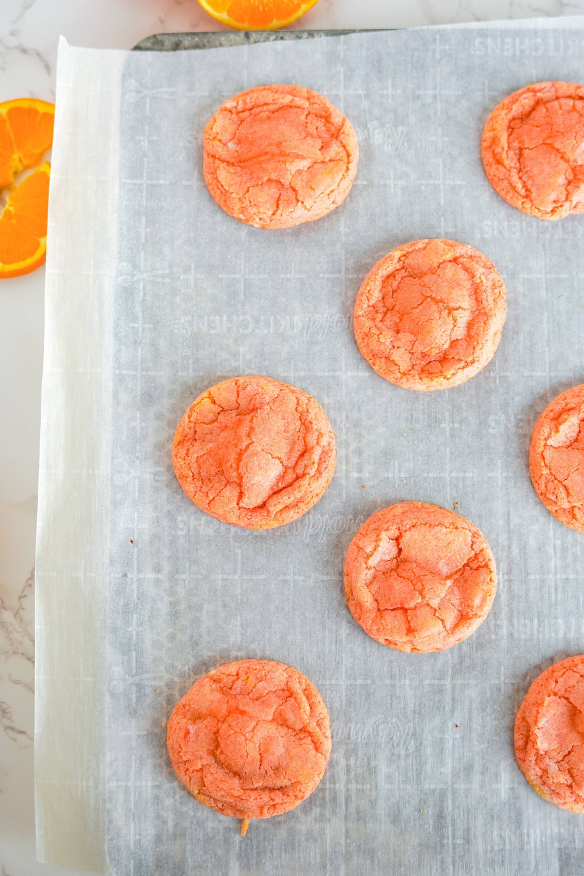 Round Orange Creamsicle Cookies are arranged on a parchment-lined baking sheet, with slices of fresh orange visible in the upper left corner.