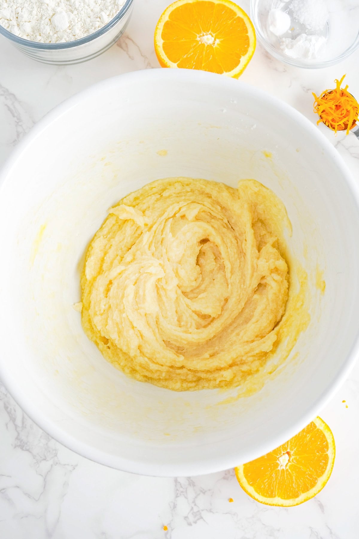 A white mixing bowl contains a creamy, blended batter for Creamsicle Cookies. Sliced oranges, zest, and bowls with flour and sugar are visible on the marble countertop.