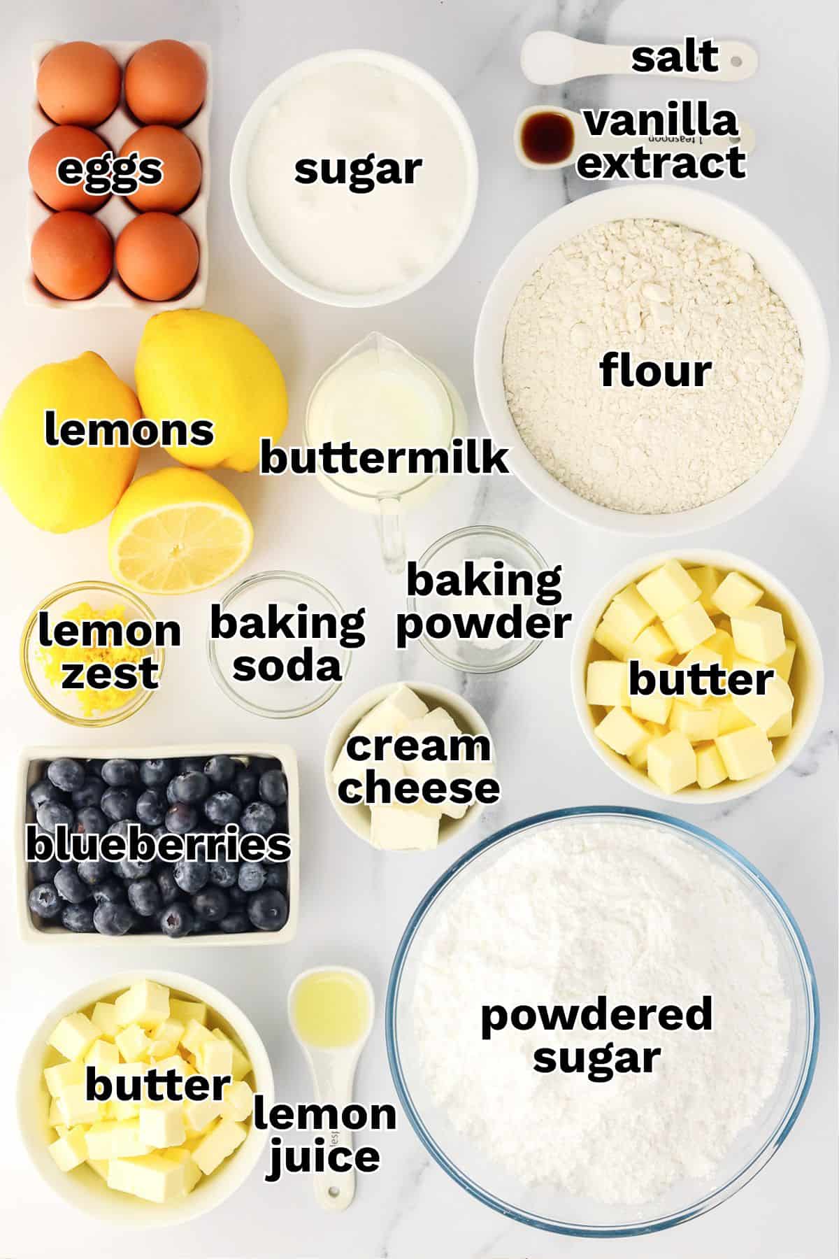 Top-down view of baking ingredients for Blueberry Lemon Cake, including eggs, sugar, flour, vanilla extract, lemons, buttermilk, baking powder, cream cheese, butter, blueberries, and powdered sugar.