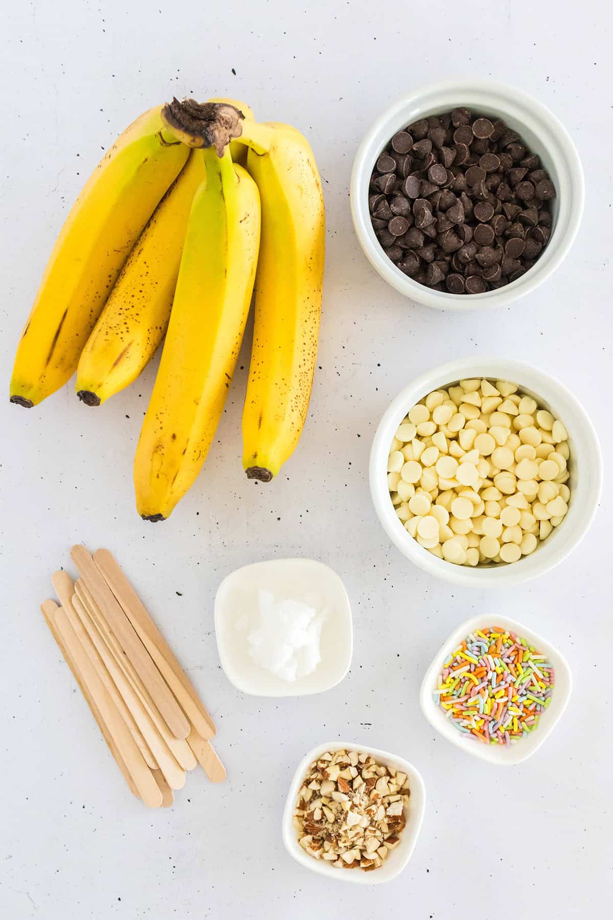 Bananas, chocolate chips, white chocolate chips, wooden sticks, coconut oil, chopped nuts, and rainbow sprinkles are arranged on a white surface for making delicious Frozen Chocolate Covered Bananas.