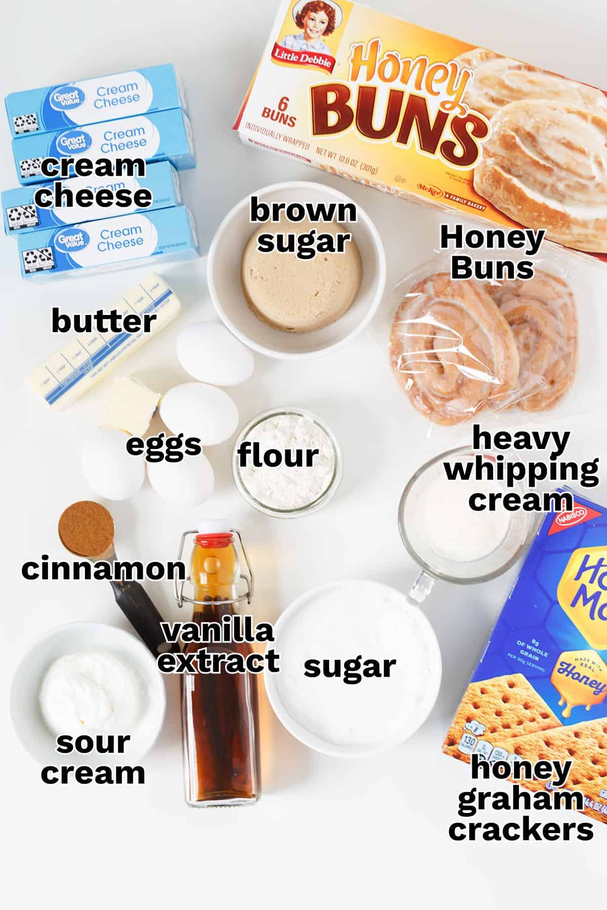 Ingredients for a Honey Bun Cheesecake recipe arranged on a white surface, including cream cheese, butter, eggs, cinnamon, sour cream, vanilla extract, brown sugar, sugar, flour, heavy cream, honey buns, and honey graham crackers.