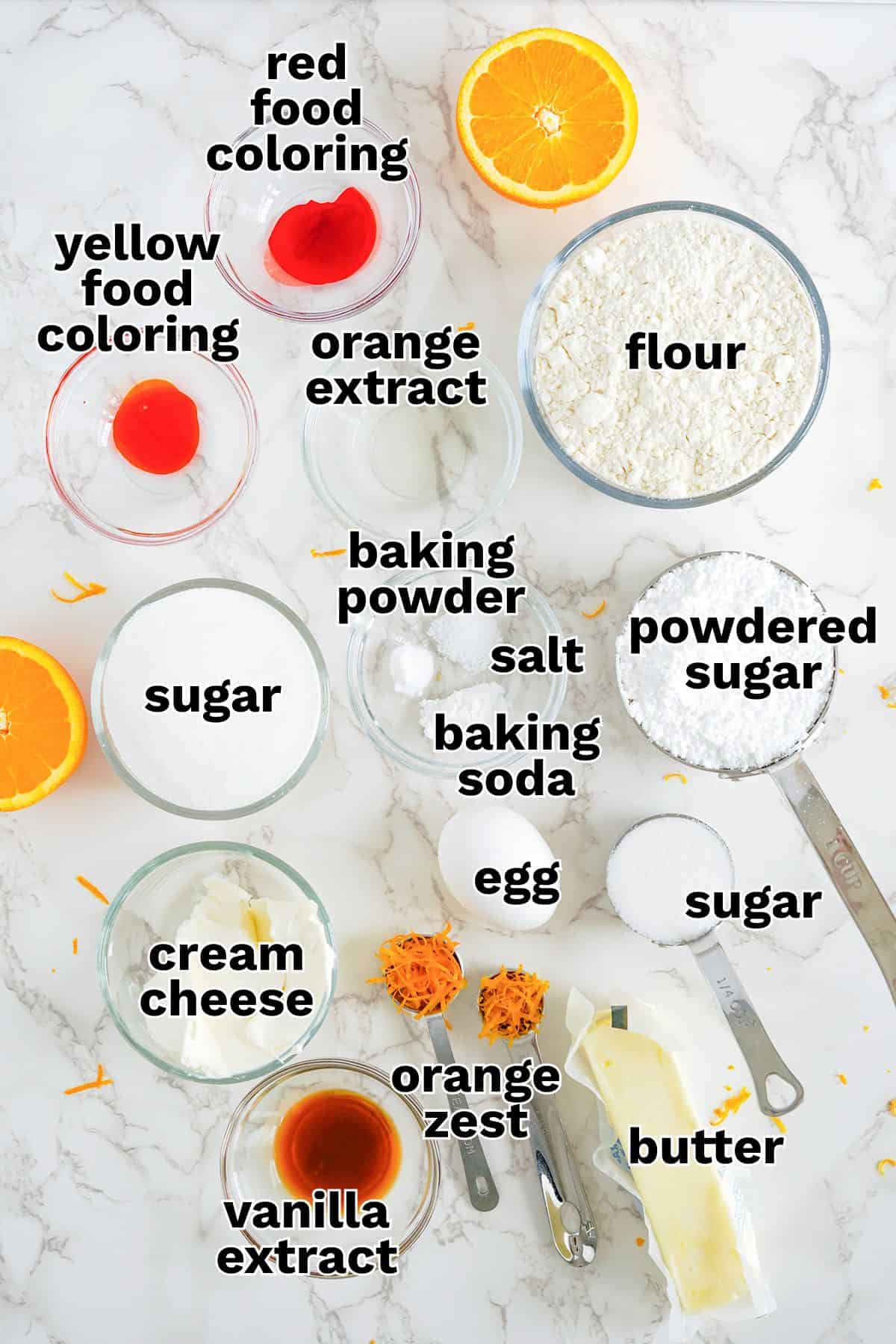 Top-down view of various labeled baking ingredients used for Orange Creamsicle Cookies, including flour, sugars, cream cheese, butter, egg, orange, extracts, food coloring, baking powder, baking soda, salt, and orange zest.