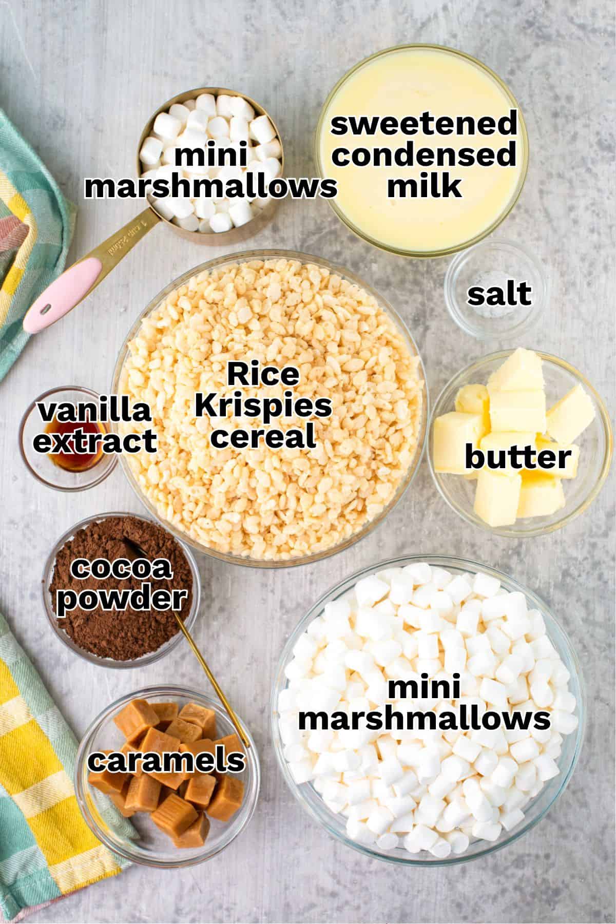 Ingredients for Star Crunch Cookies arranged on a table: mini marshmallows, sweetened condensed milk, salt, butter, Rice Krispies cereal, vanilla extract, cocoa powder, and caramels.