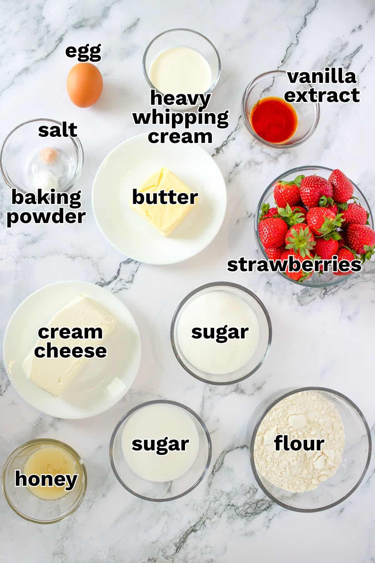 Ingredients for strawberry pizza arranged on a marble surface, including an egg, butter, heavy whipping cream, vanilla extract, strawberries, cream cheese, sugar, flour, honey, baking powder, and salt.