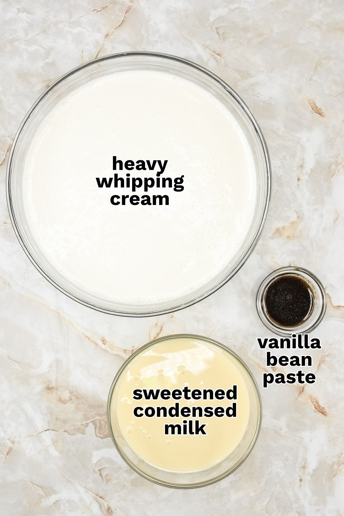 Three labeled ingredients for no churn vanilla ice cream on a marble surface: a bowl of heavy whipping cream, a bowl of sweetened condensed milk, and a small dish of vanilla bean paste.