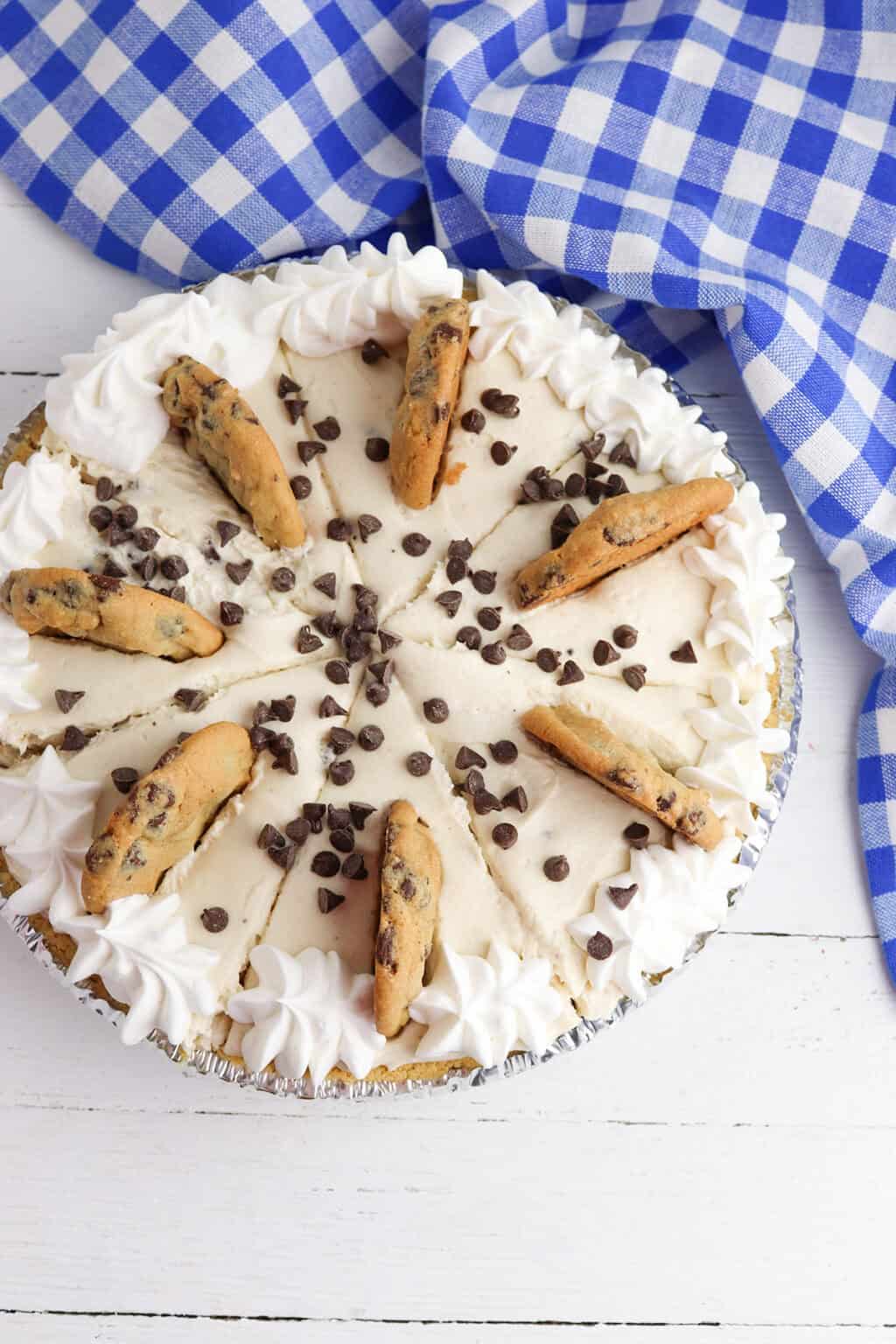 A Cookie Dough Cheesecake Pie topped with whipped cream, chocolate chip cookies, and chocolate chips sits on a white wooden surface next to a blue and white checkered cloth.