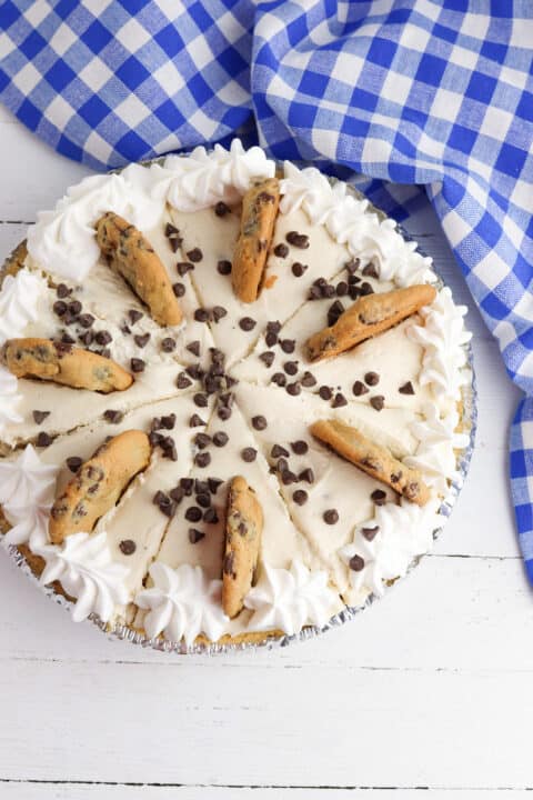 A Cookie Dough Cheesecake Pie topped with whipped cream, chocolate chip cookies, and chocolate chips sits on a white wooden surface next to a blue and white checkered cloth.