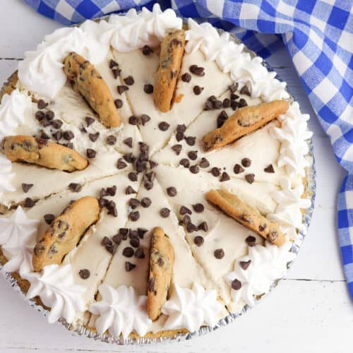 A Cookie Dough Cheesecake Pie topped with whipped cream, chocolate chip cookies, and chocolate chips sits on a white wooden surface next to a blue and white checkered cloth.
