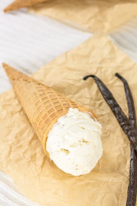 A scoop of creamy vanilla ice cream in a waffle cone rests on parchment paper next to two dried vanilla beans, perfect for indulging in a classic no churn treat.