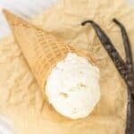 A scoop of no churn vanilla ice cream in a waffle cone is placed on crumpled parchment paper next to two vanilla beans.
