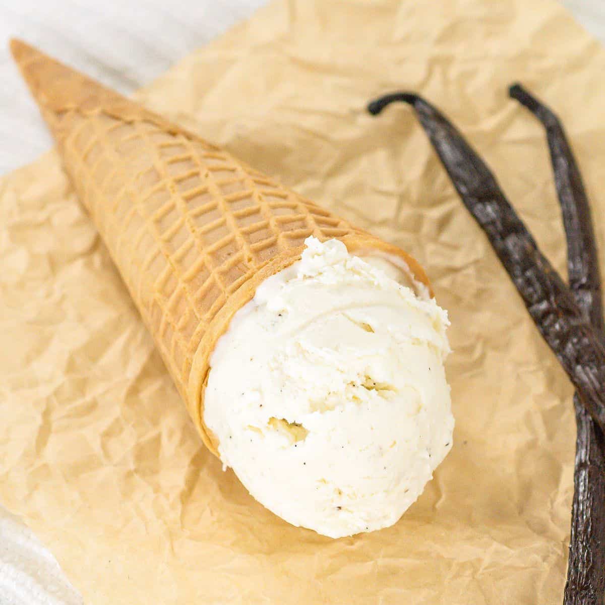 A scoop of no churn vanilla ice cream in a waffle cone is placed on crumpled parchment paper next to two vanilla beans.