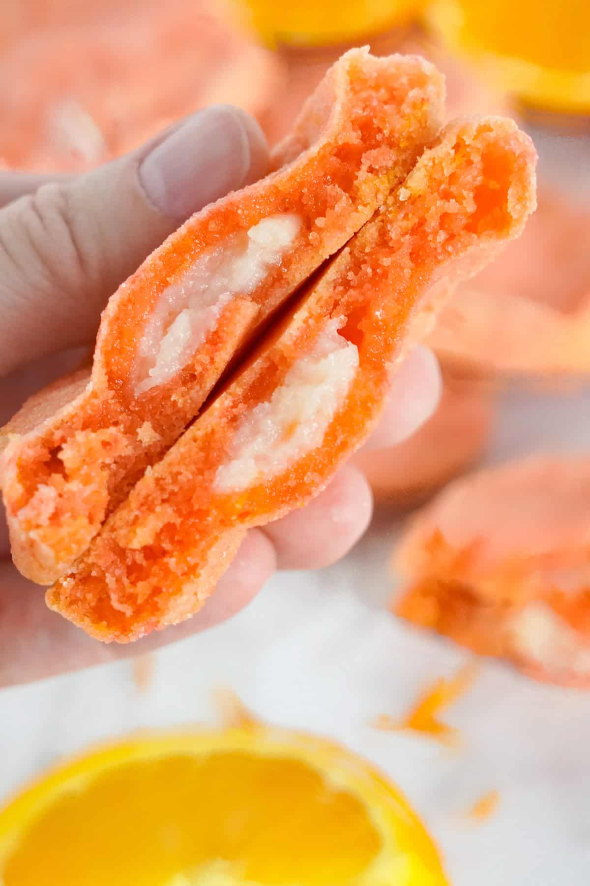 A hand holding a split Orange Creamsicle Cookie with a creamy white filling, with more cookies and an orange slice in the background.