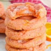 A stack of five Orange Creamsicle Cookies with a bite taken from the top cookie, surrounded by orange slices on a marble surface.