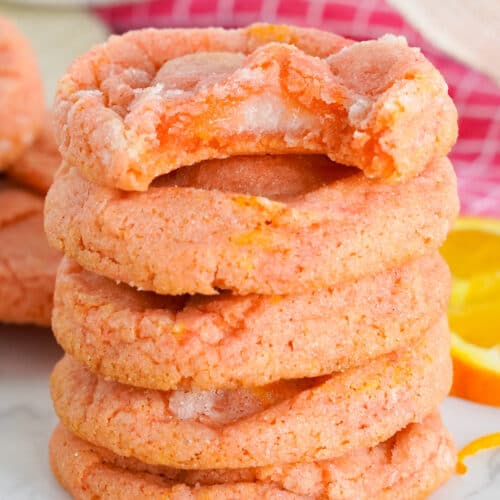A stack of five Orange Creamsicle Cookies with a bite taken from the top cookie, surrounded by orange slices on a marble surface.