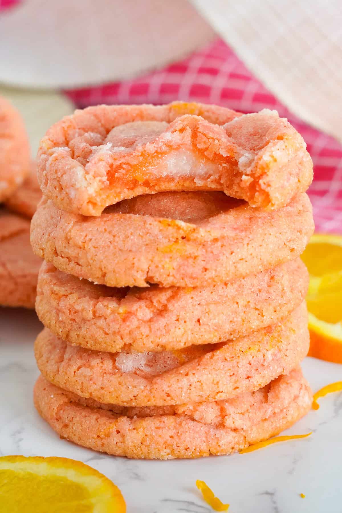 A stack of five Orange Creamsicle Cookies with a bite taken from the top cookie, surrounded by orange slices on a marble surface.