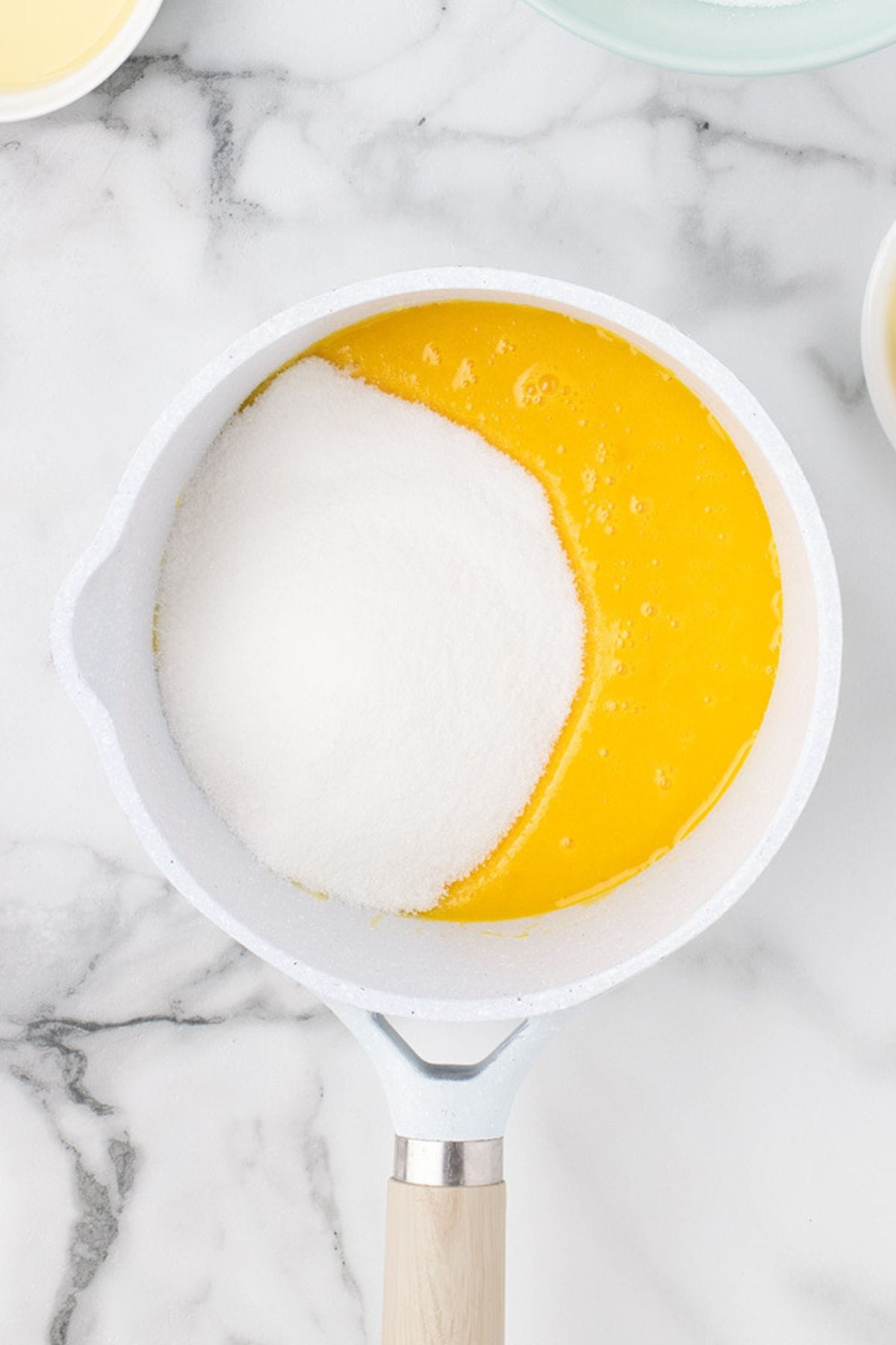 A white saucepan containing egg yolks and granulated sugar sits on a marble countertop.