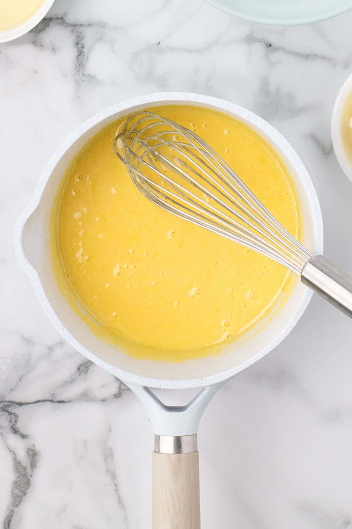 A metal whisk rests in a white saucepan filled with a yellow liquid mixture, placed on a marble countertop.