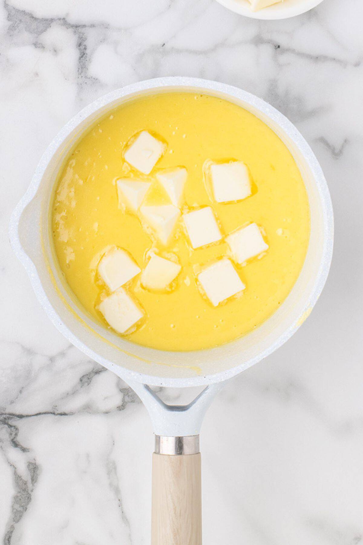 A saucepan on a marble surface contains a yellow liquid mixture with several cubes of butter melting on top.