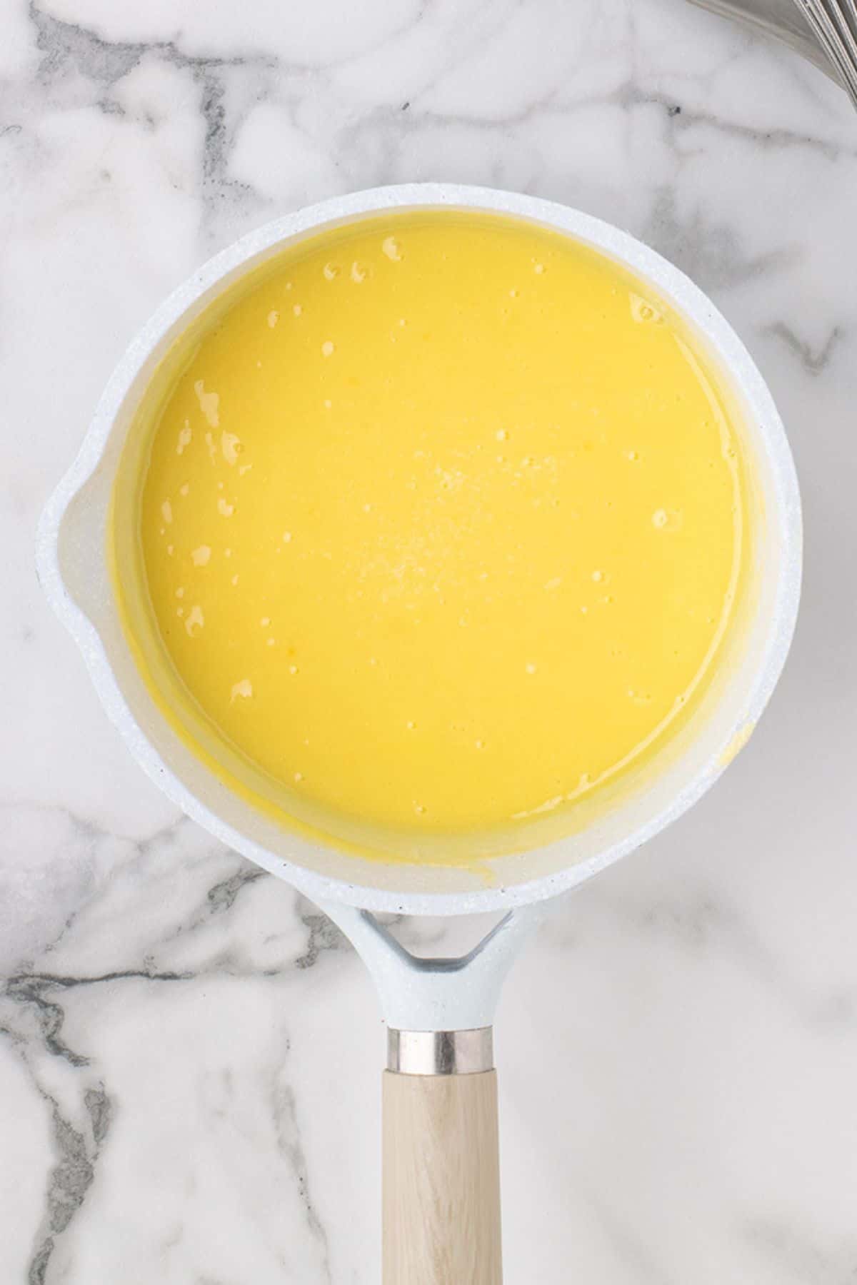 A white saucepan filled with a smooth yellow liquid, placed on a marble countertop.