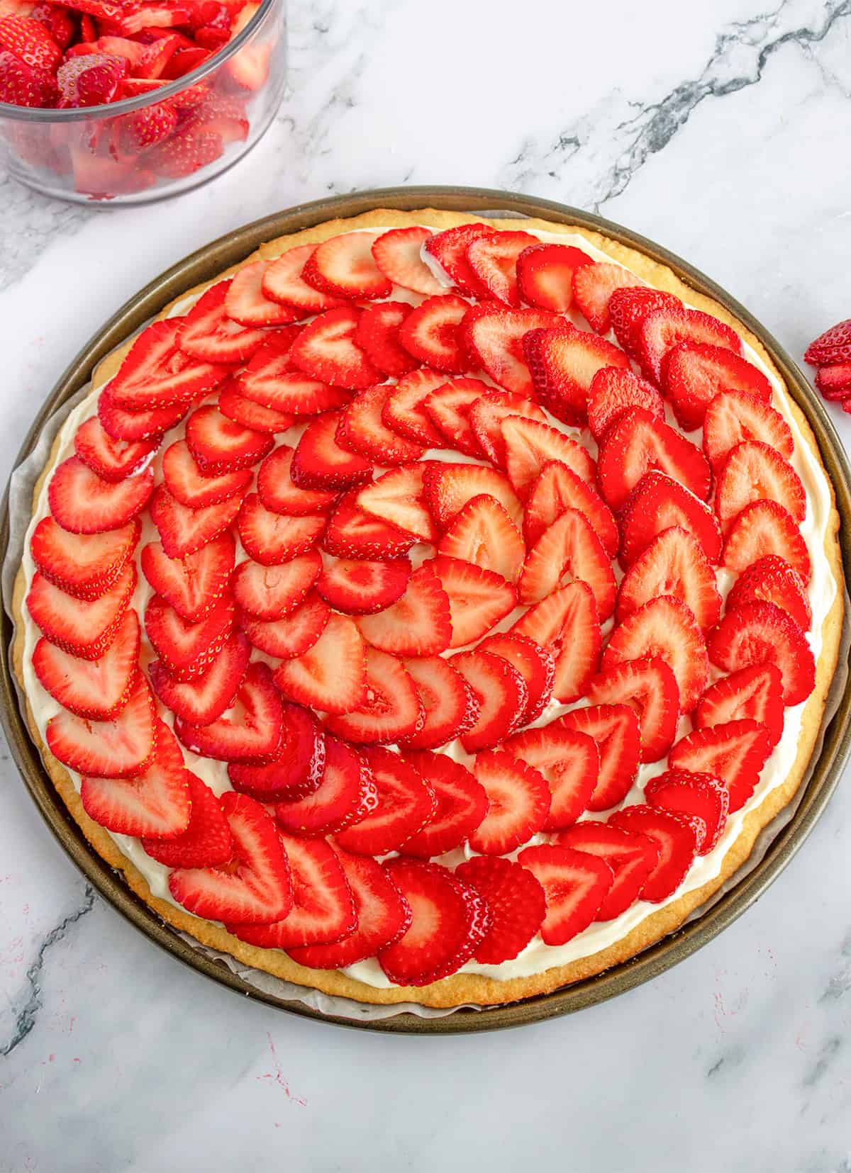 A delightful Strawberry Pizza featuring a spiral pattern of fresh strawberry slices on a creamy white base, beautifully displayed on a round baking tray.
