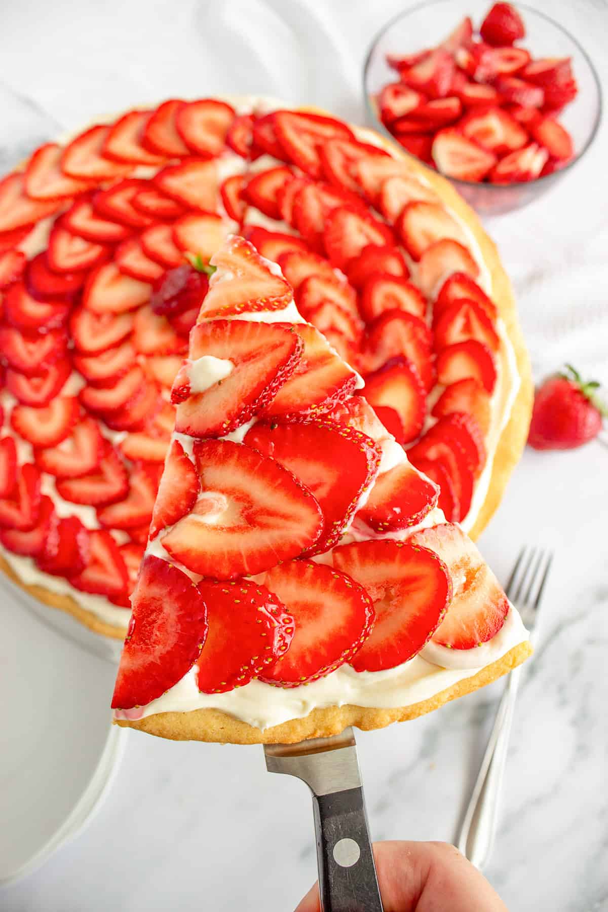 A hand holds a slice of strawberry dessert pizza with cream filling, topped with fresh sliced strawberries. The whole tart and a bowl of extra strawberries are in the background.